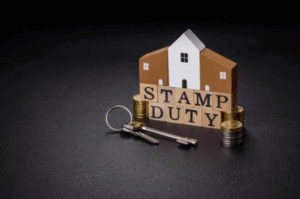 House model with wooden blocks spelling ‘Stamp Duty’, surrounded by coins and house keys, symbolising property purchase costs and stamp duty obligations.
