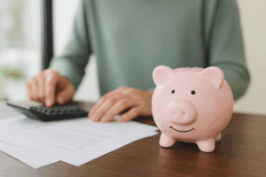 Person calculating finances with a pink piggy bank on a desk, representing how genuine savings help first home buyers qualify for a home loan.