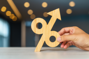 Hand holding a wooden percentage symbol with upward arrow, representing rising interest rates and lending trends against a blurred office background.