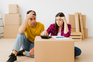 Young couple sitting among moving boxes calculating home buying costs, stamp duty, deposit requirements, and upfront fees using a calculator and paperwork.